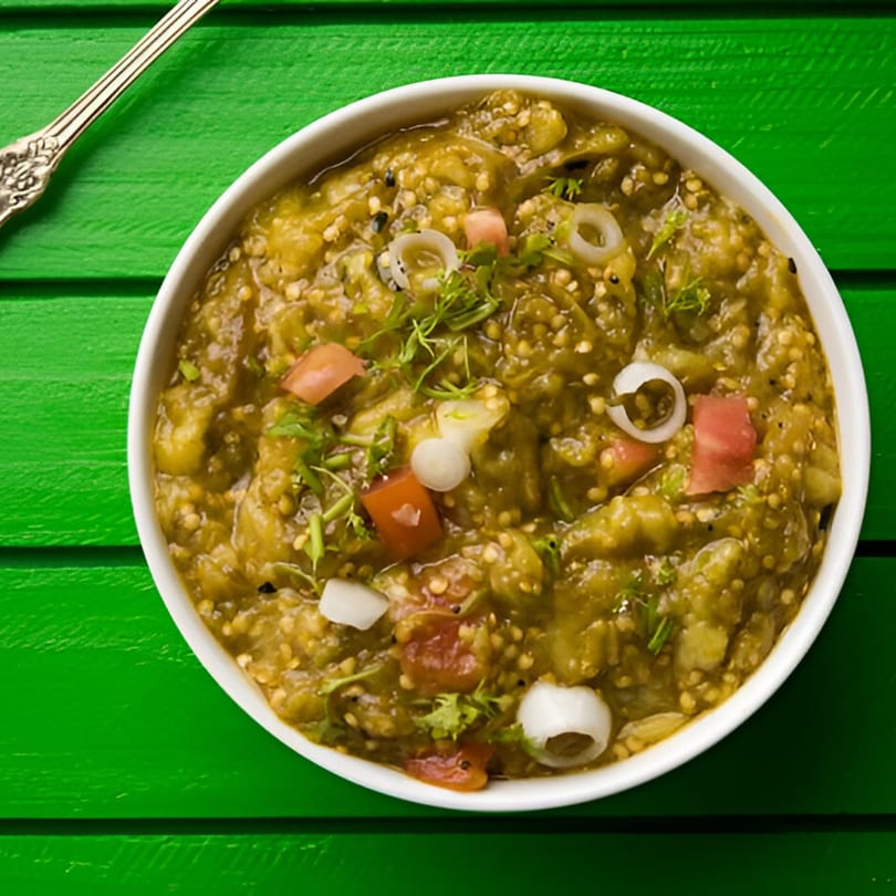 A white bowl of green Indian baingan bharta, garnished with chopped tomatoes, onions, and herbs.