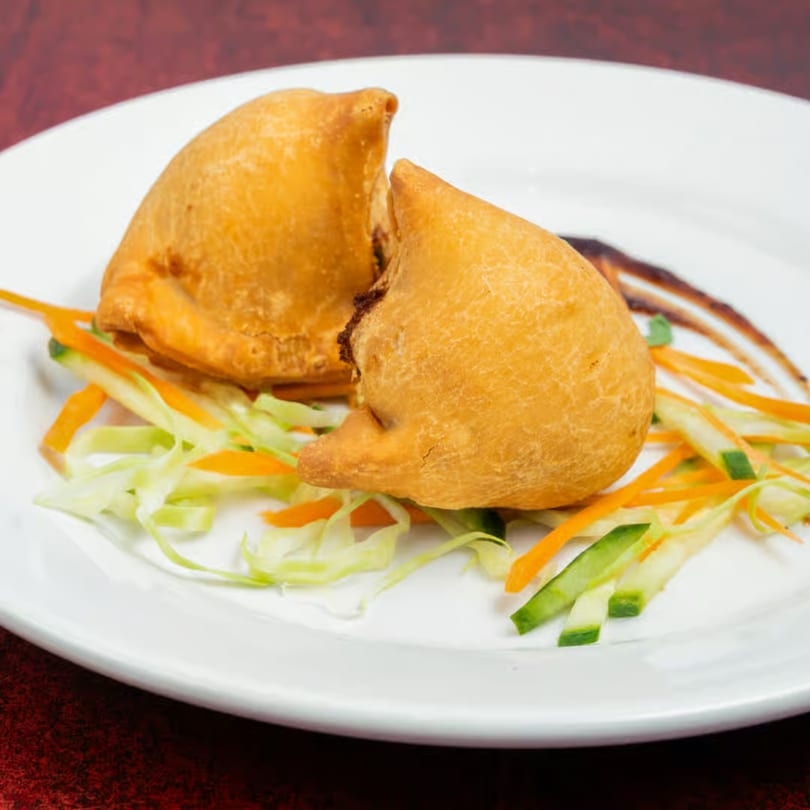 Two golden-brown samosas on a white plate, garnished with shredded carrots, cabbage, and a dark sauce.